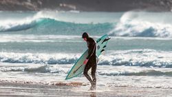 Surfer in a wetsuit carrying a surfboard on a beach with waves in the background