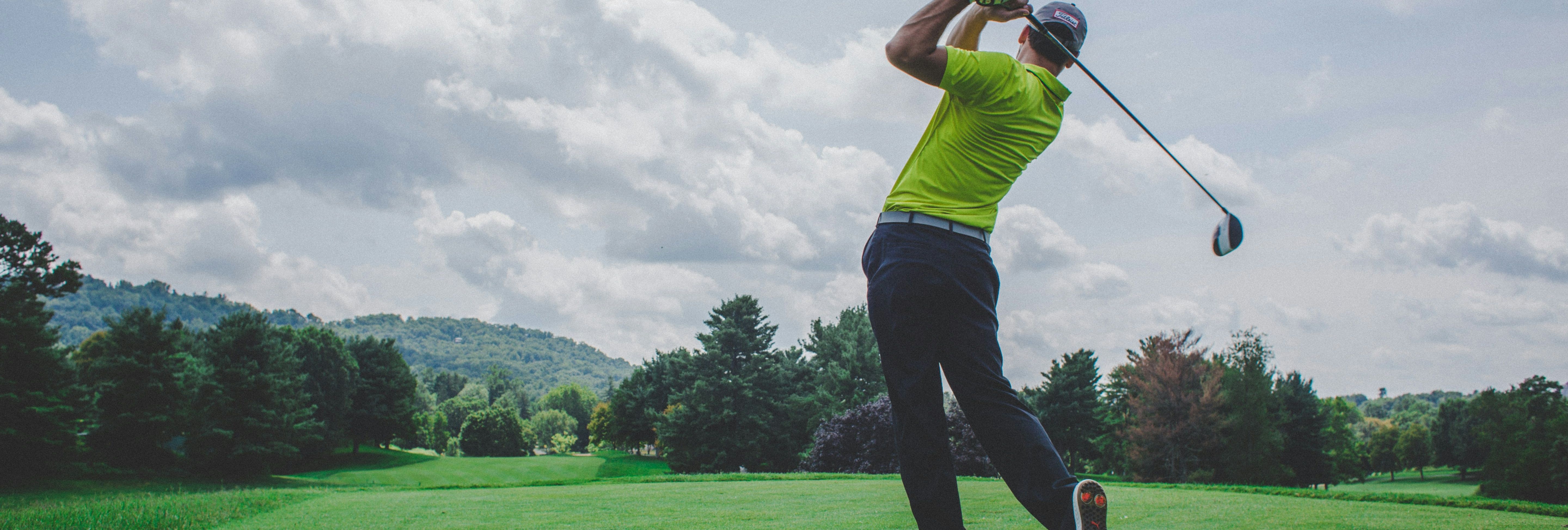 Golfer in a yellow shirt taking a swing on a lush golf course with trees and hills in the background