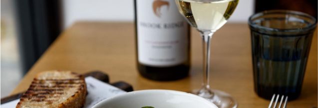 A bowl of gourmet sardines served with greens and caviar, accompanied by toasted bread, a glass of white wine, and cutlery on a wooden table.