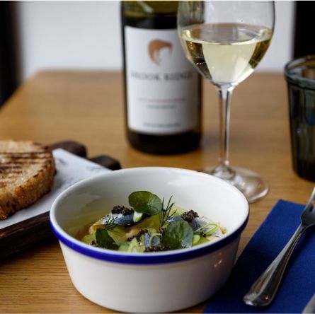 A bowl of gourmet sardines served with greens and caviar, accompanied by toasted bread, a glass of white wine, and cutlery on a wooden table.