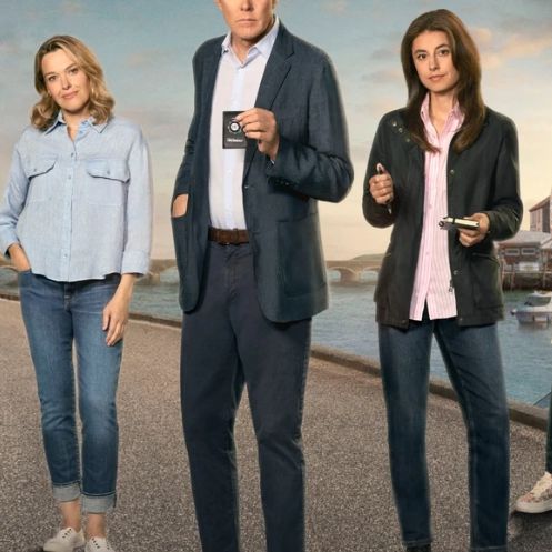 Five people standing on a coastal street in front of waterfront buildings and boats, with a duck in the foreground.