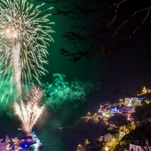 Fireworks display over a coastal town at night with illuminated houses and green smoke in the sky.