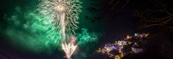 Fireworks display over a coastal town at night with illuminated houses and green smoke in the sky.
