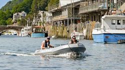 Two people riding an inflatable boat on a river with boats and buildings along the shore.