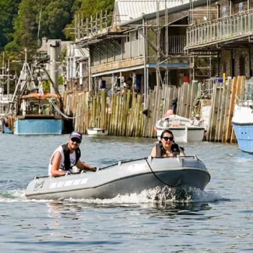 Two people riding an inflatable boat on a river with boats and buildings along the shore.