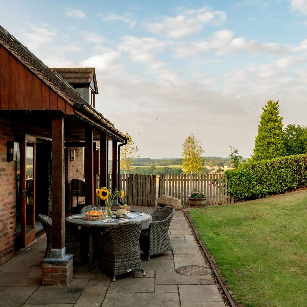 Outdoor patio area with a round table and chairs beside a brick house, overlooking a fenced yard with trees and hedges.