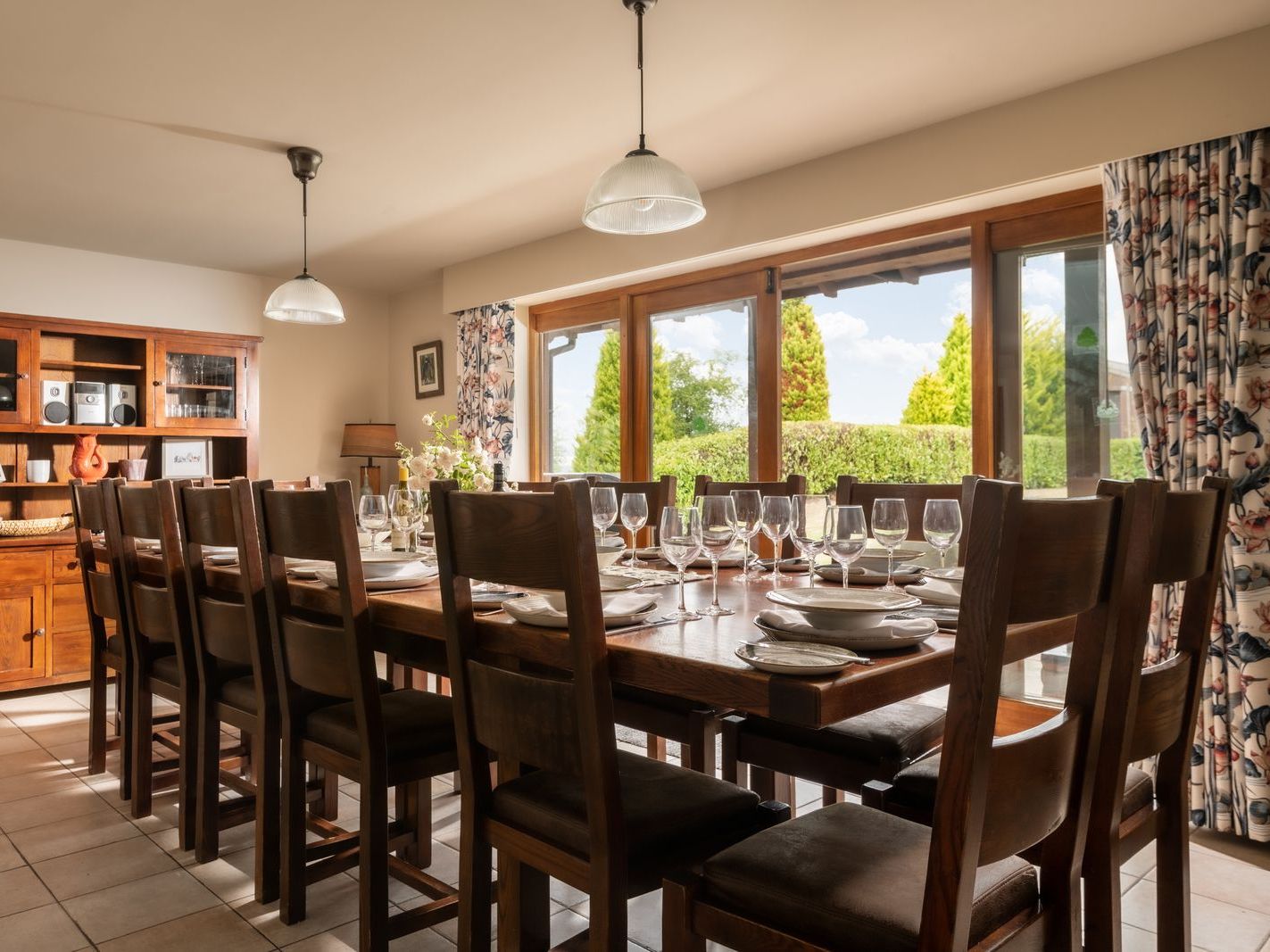 Elegant dining room with a long wooden table set for a meal, surrounded by chairs and large windows overlooking a garden.