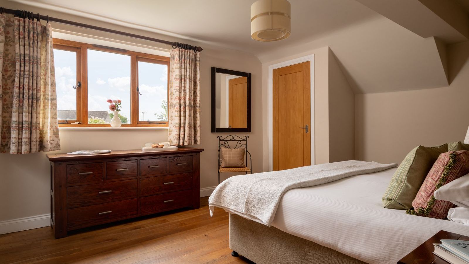 Cozy bedroom with wooden furniture, bed with pillows, window with floral curtains, dresser, and chair.
