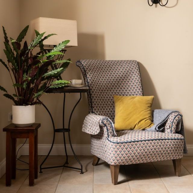 Cozy reading nook with patterned armchair, yellow pillow, side tables, indoor plant, lamp, and wall sconce.