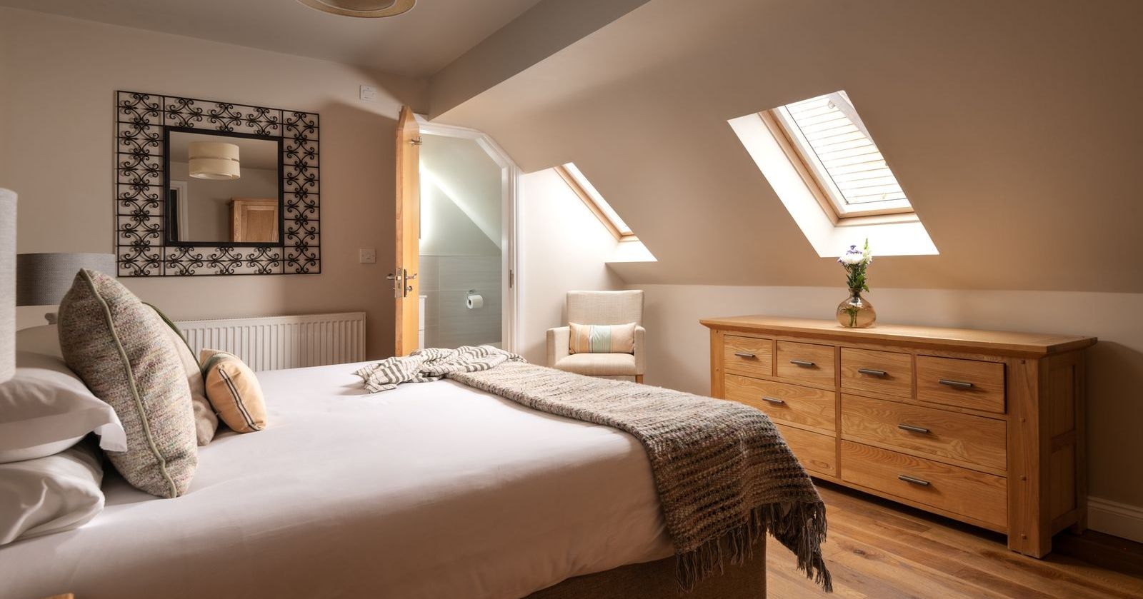 Cozy attic bedroom with sloped ceiling, skylight windows, a bed with pillows and blanket, wooden dresser, and armchair