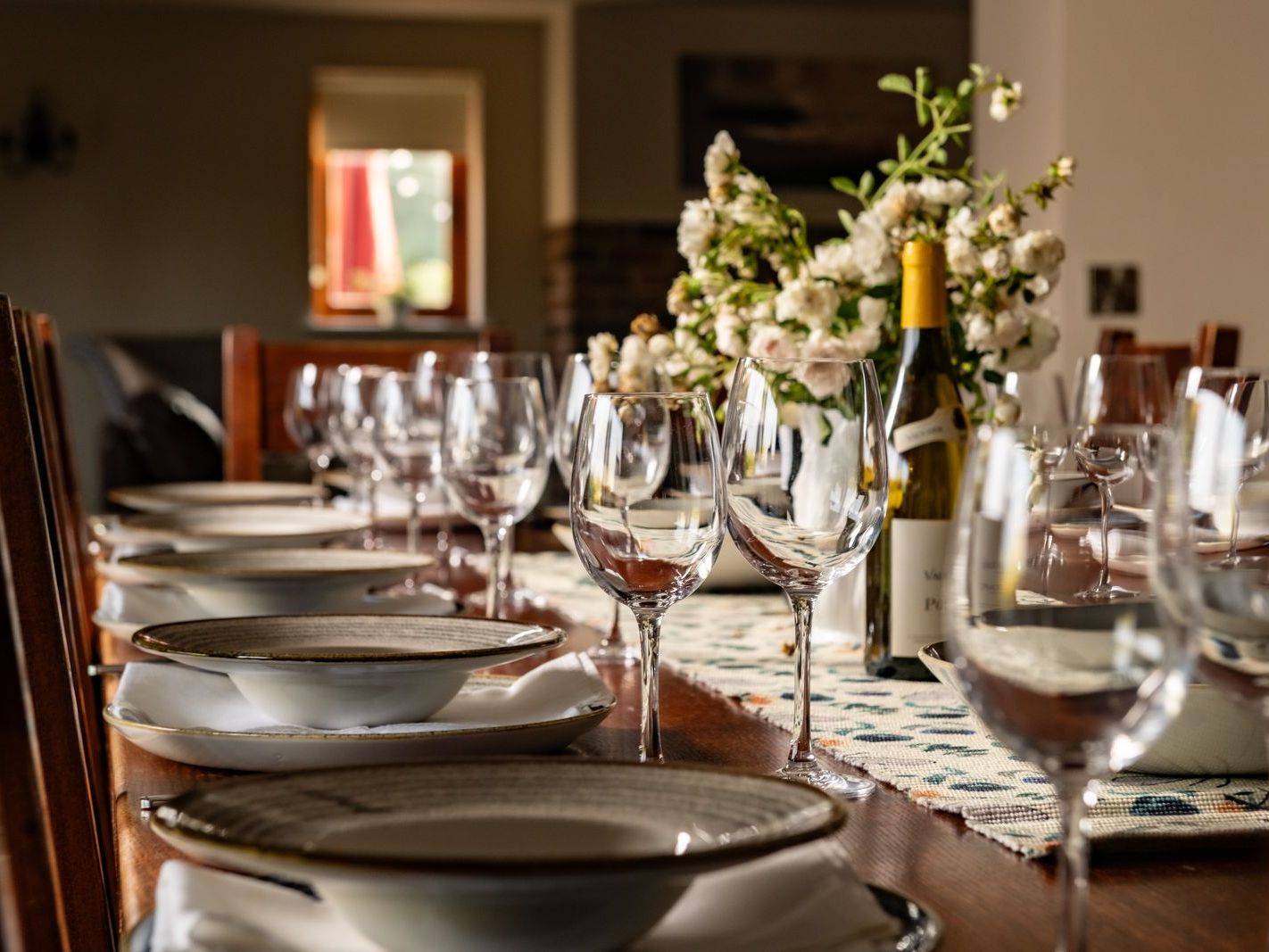 Elegant dining table set with glassware, plates, napkins, flowers, and a bottle of wine