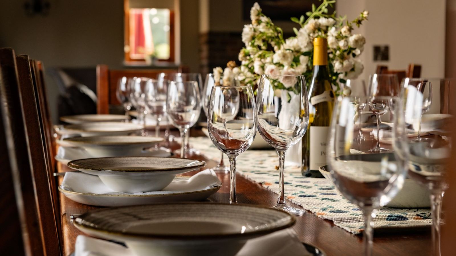 Elegant dining table set with glassware, plates, napkins, flowers, and a bottle of wine