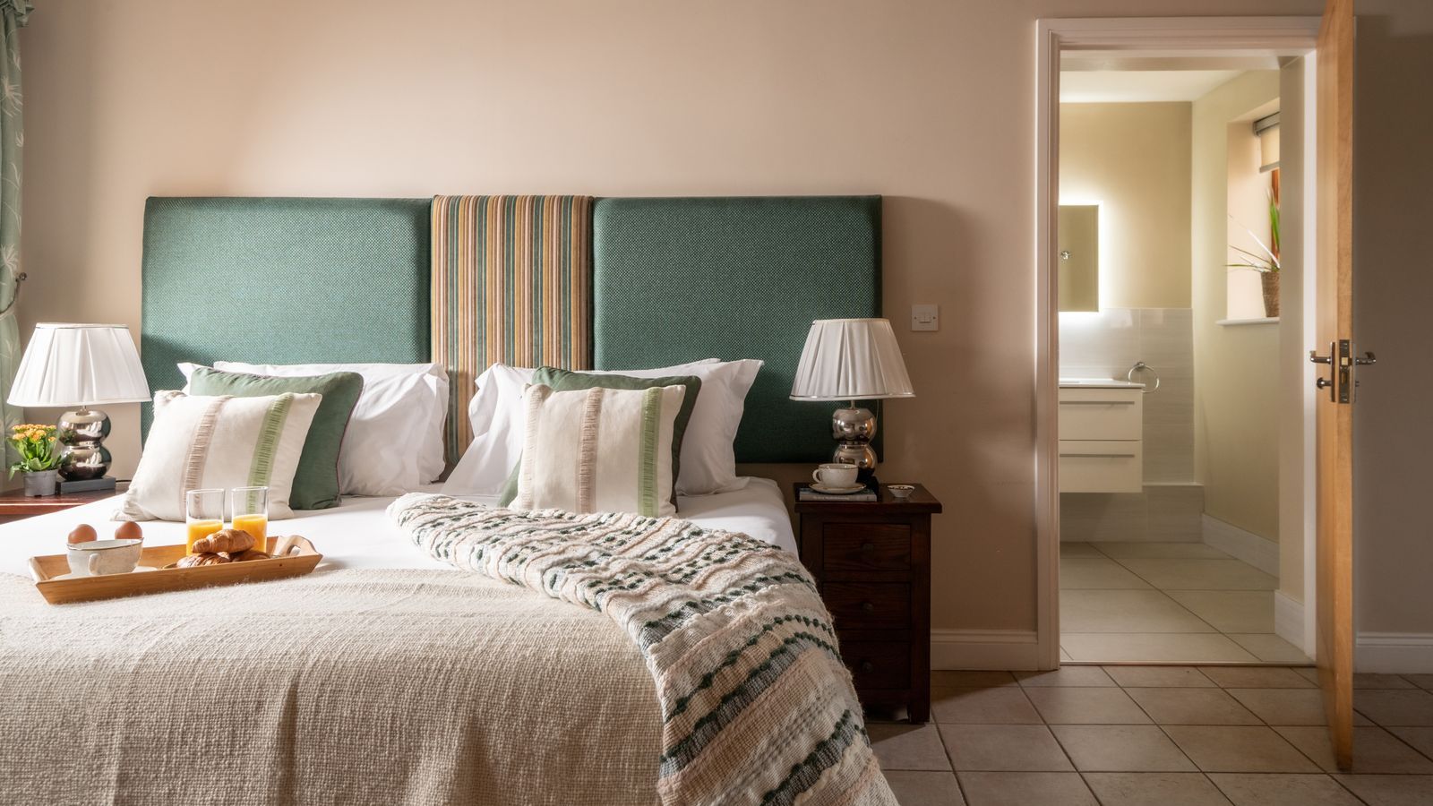 Cozy bedroom with a large bed, green upholstered headboard, bedside tables with lamps, and a view into an ensuite bathroom.