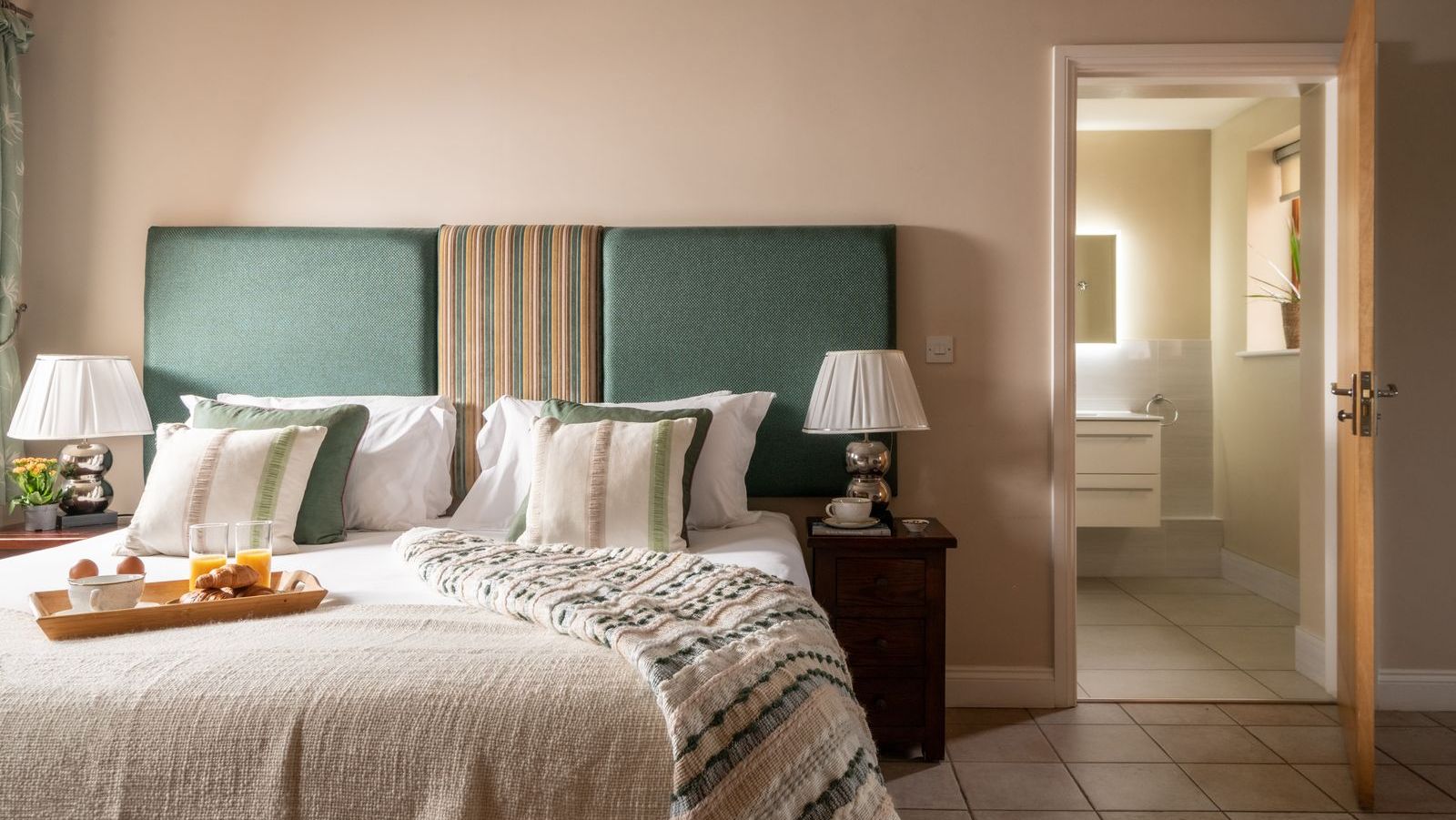 Cozy bedroom with a large bed, green upholstered headboard, bedside tables with lamps, and a view into an ensuite bathroom.