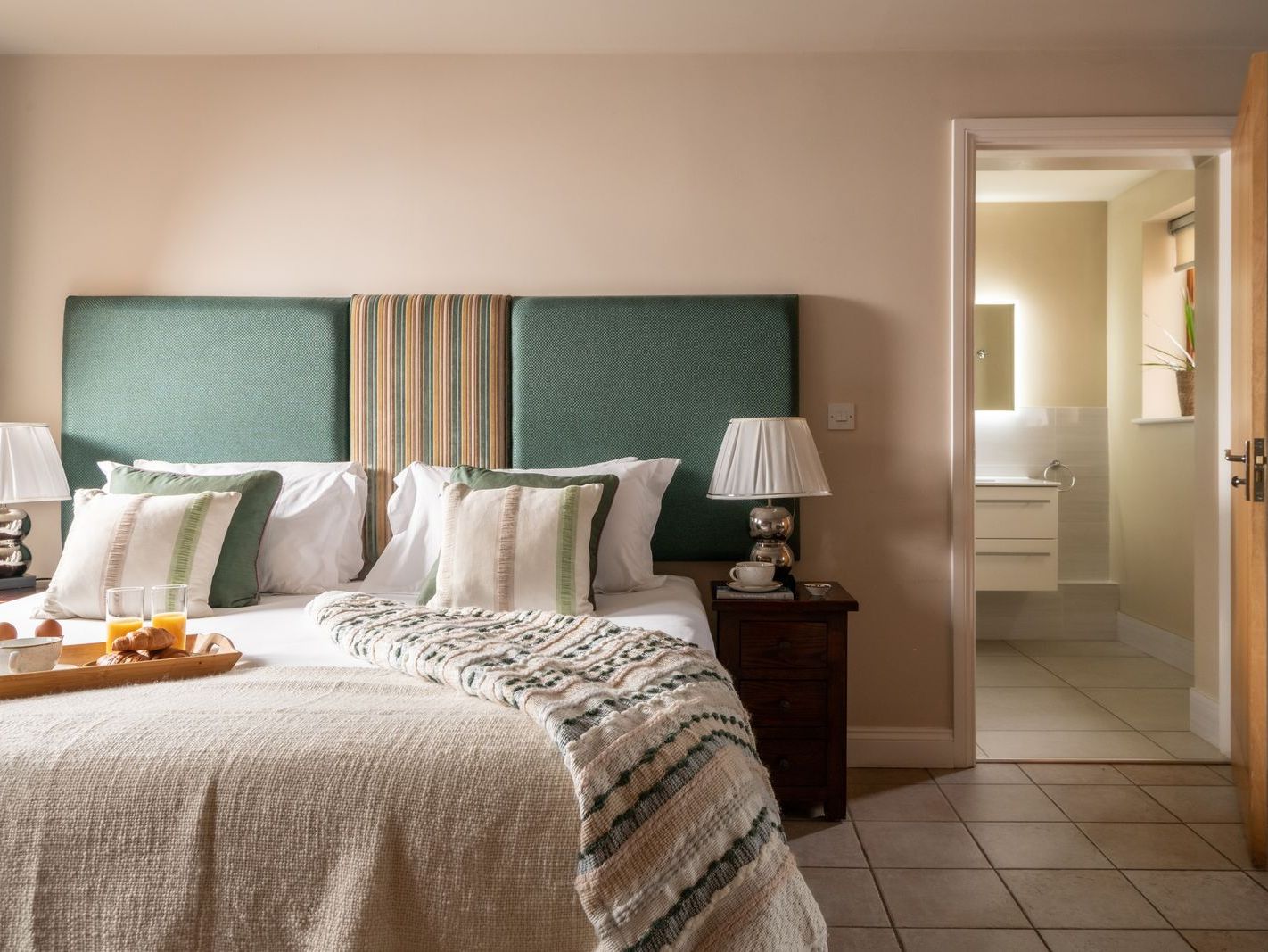 Cozy bedroom with a large bed, green upholstered headboard, bedside tables with lamps, and a view into an ensuite bathroom.