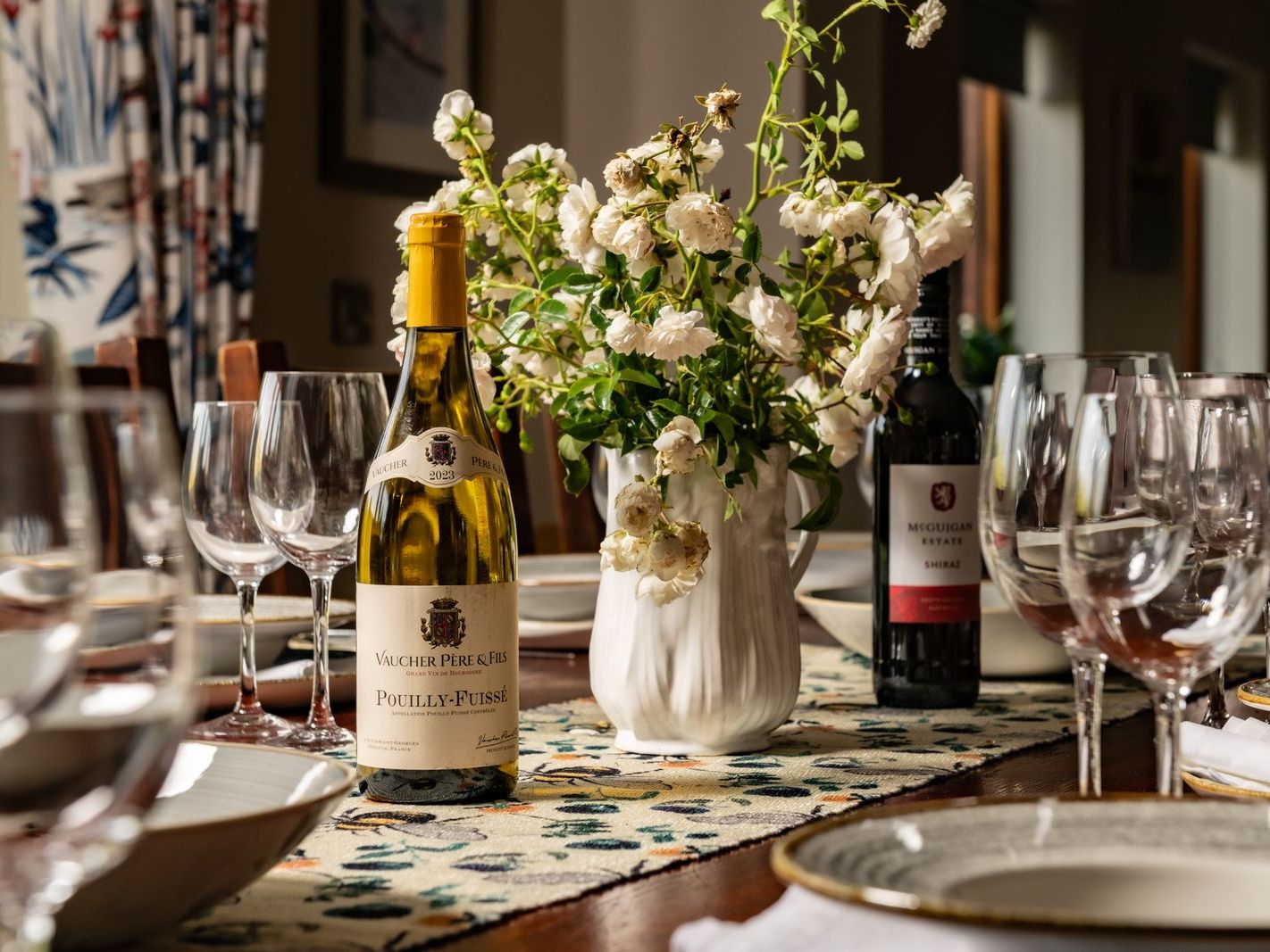 Elegant dining table set with wine bottles, glassware, bowls, and a vase of white flowers.