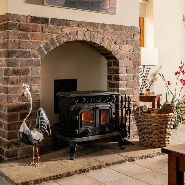 Cozy living room fireplace with a wood stove set in a brick alcove, a wicker basket of firewood, and a decorative crane sculpture.