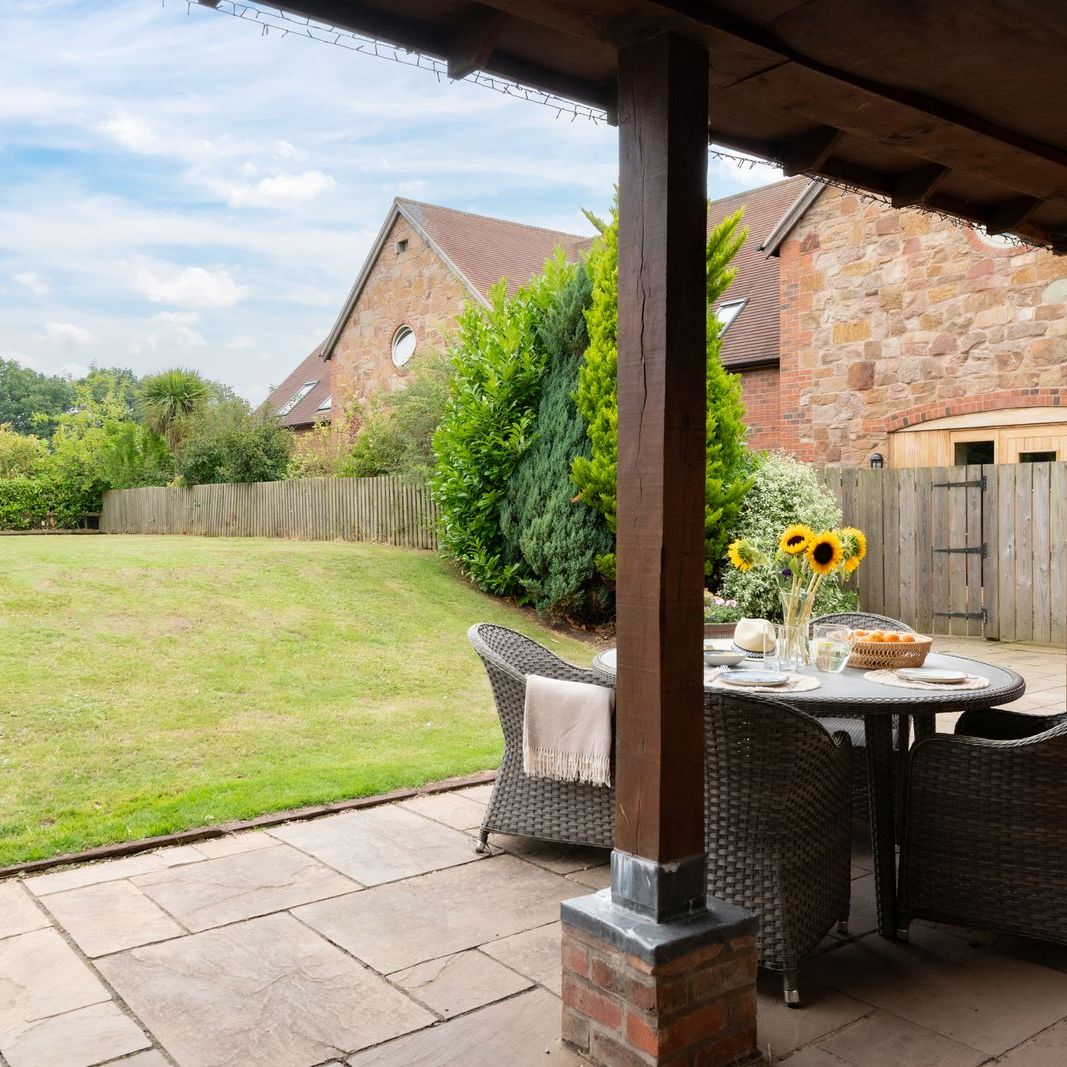Covered patio area with a round table and chairs overlooking a grassy backyard and stone house exterior.