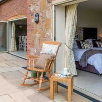 Outdoor patio area with a wooden chair and table next to a bedroom with open folding doors.