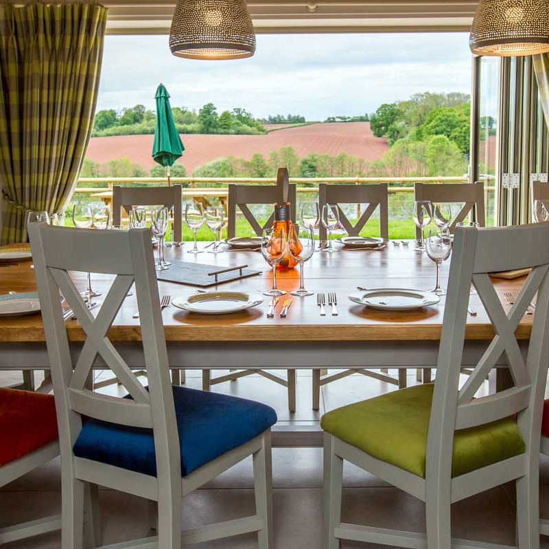 Elegant dining room with a large wooden table set for a meal, colorful chair cushions, and a scenic countryside view through open glass doors.