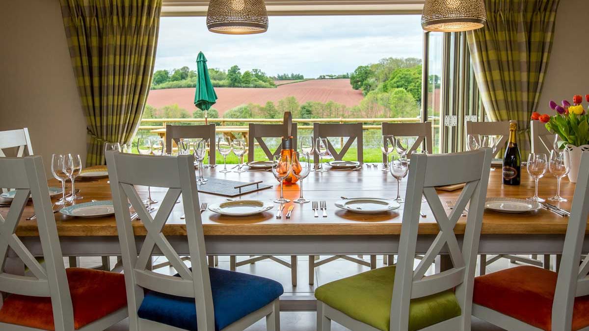 Elegant dining room with a large wooden table set for a meal, colorful chair cushions, and a scenic countryside view through open glass doors.