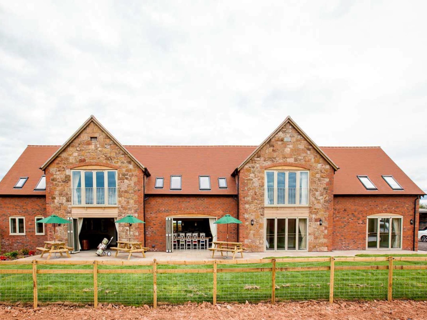 Large modern barn-style house with red brick and stone exterior, outdoor seating, and fenced lawn