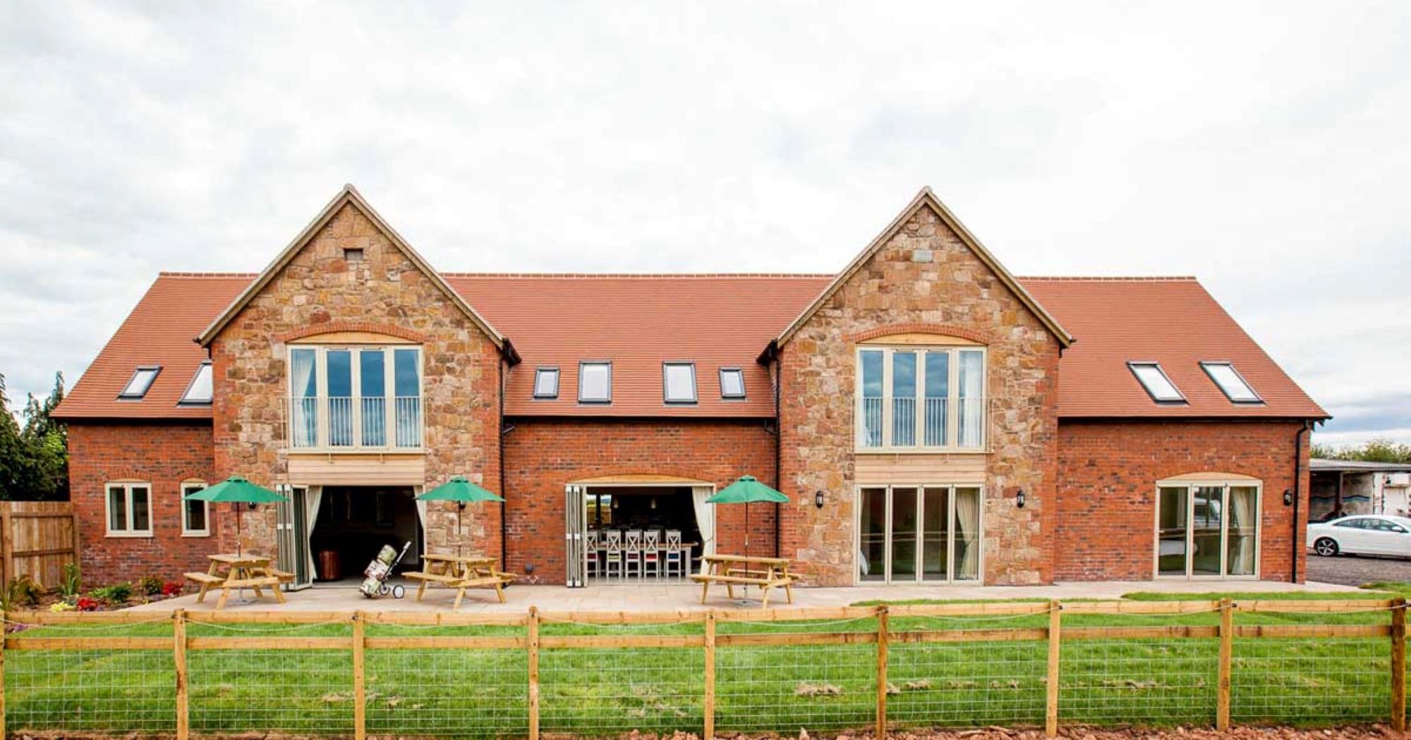 Large modern barn-style house with red brick and stone exterior, outdoor seating, and fenced lawn