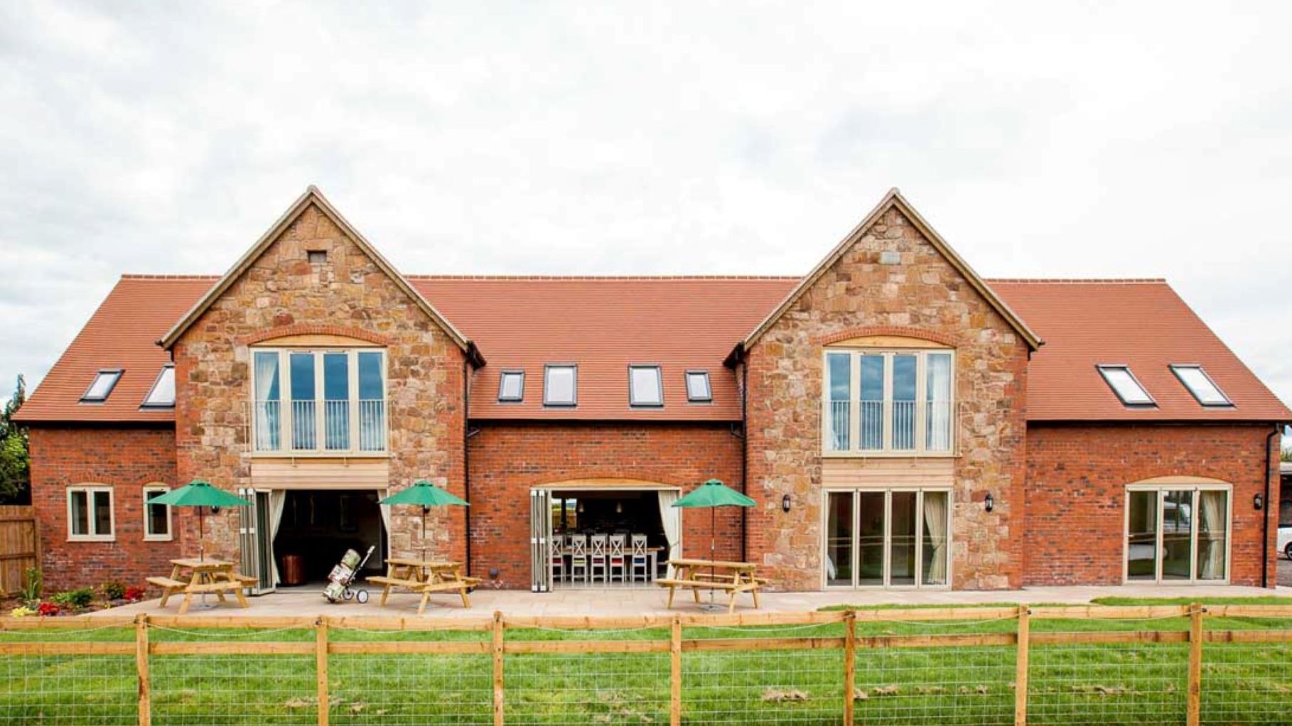 Large modern barn-style house with red brick and stone exterior, outdoor seating, and fenced lawn