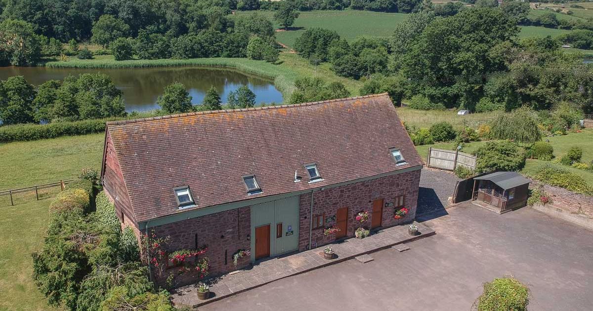 Aerial view of a rustic stone barn surrounded by countryside, trees, and a pond.