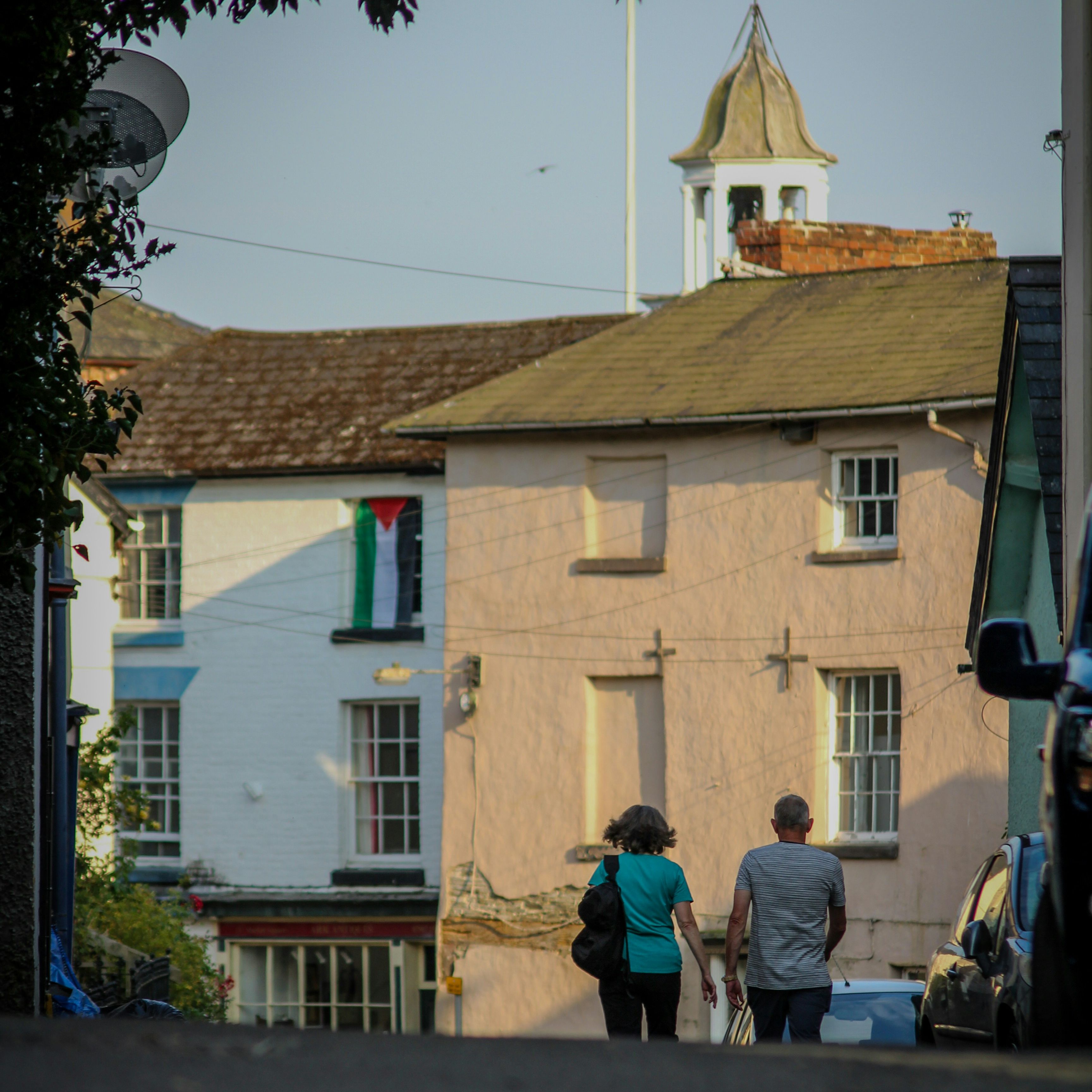 Two people walking up a quiet street in a small town with pastel-colored houses and a church steeple in the background.