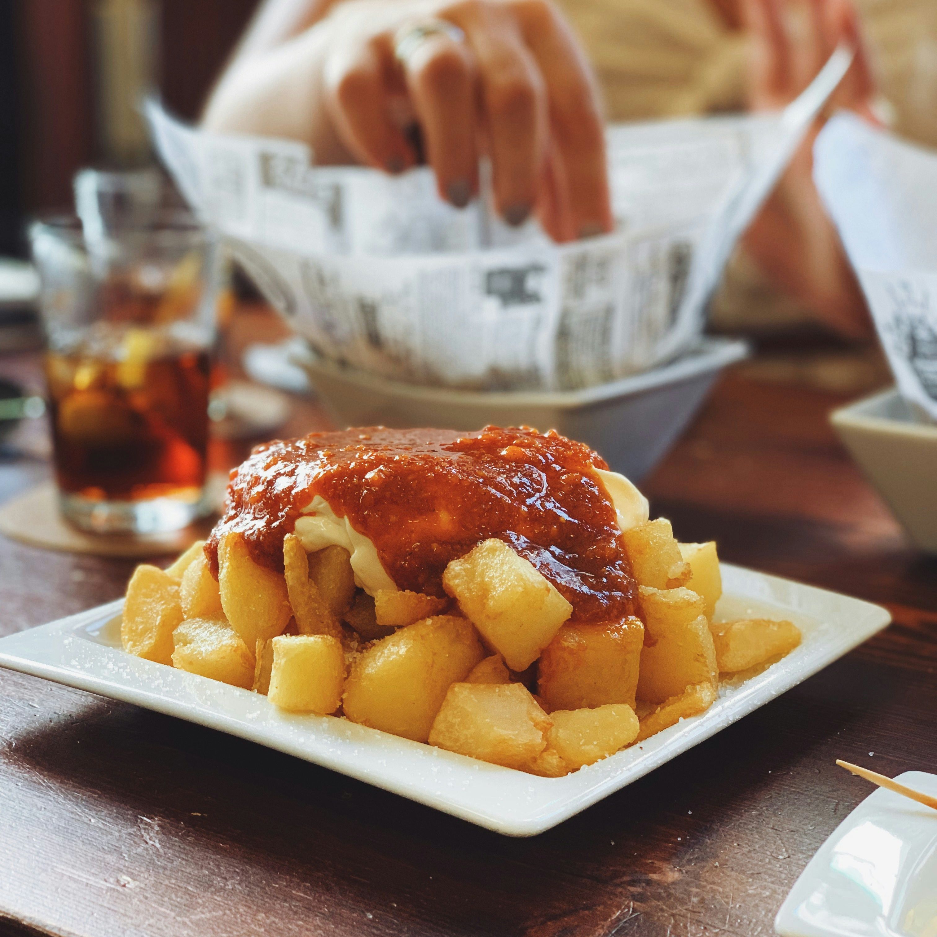 A plate of patatas bravas, Spanish fried potatoes topped with spicy tomato sauce and aioli, on a wooden table with a hand reaching for food in the background.