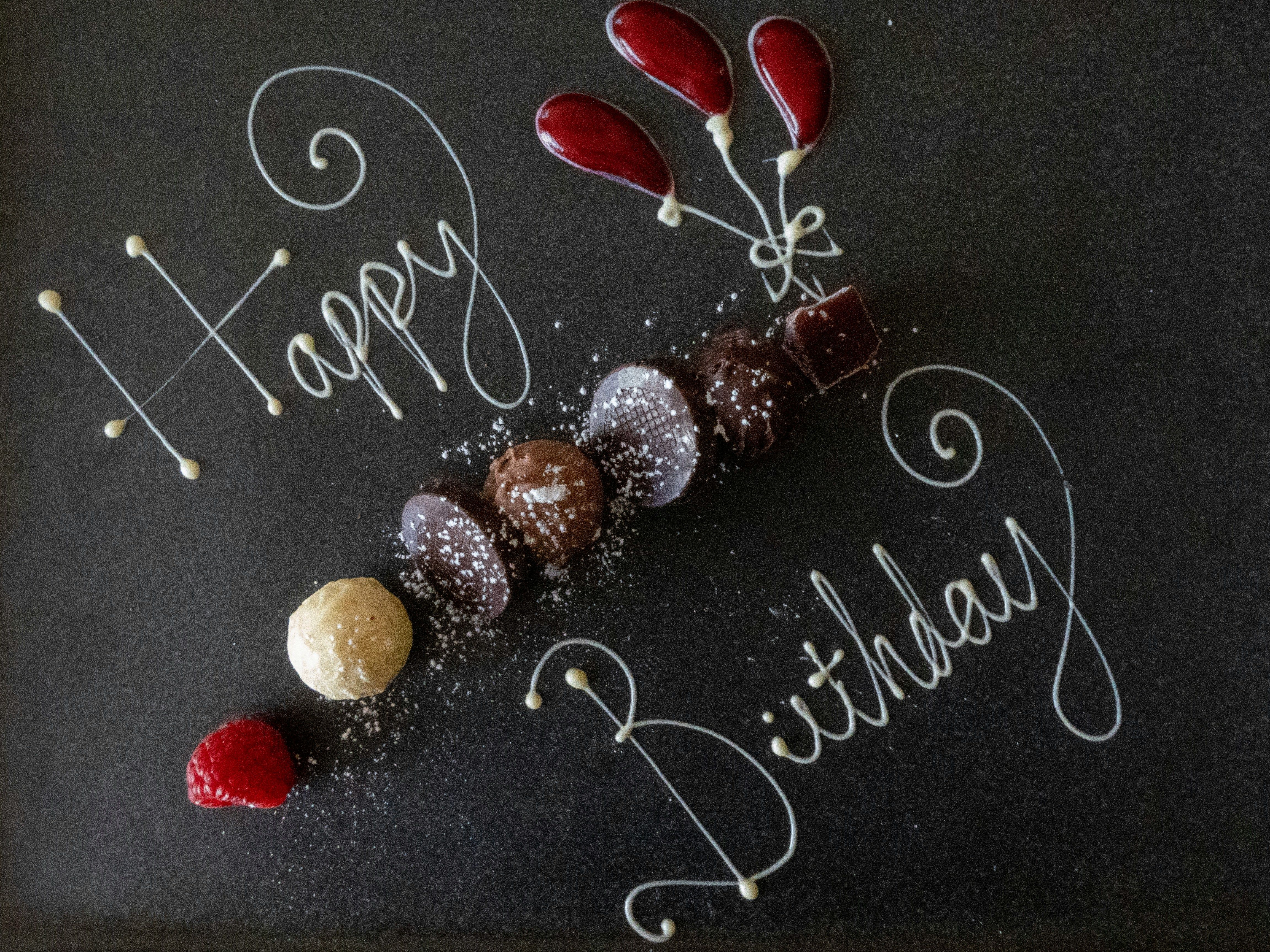 Elegant dessert plate with 'Happy Birthday' written in white chocolate, assorted chocolates, raspberry, and red sauce balloons.