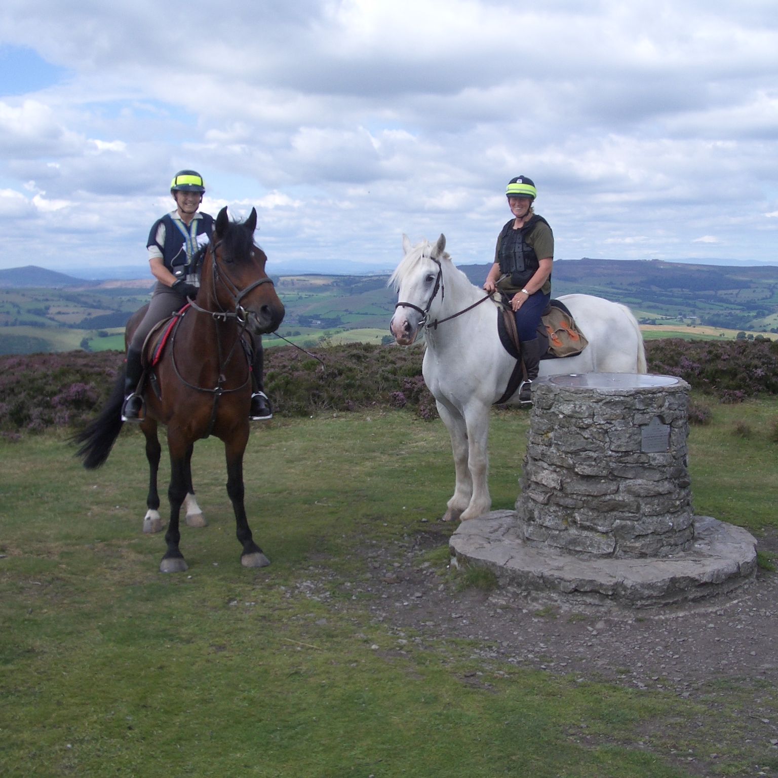 Two people on horseback next to a stone marker on a grassy hilltop, with scenic rolling hills and sky in the background.
