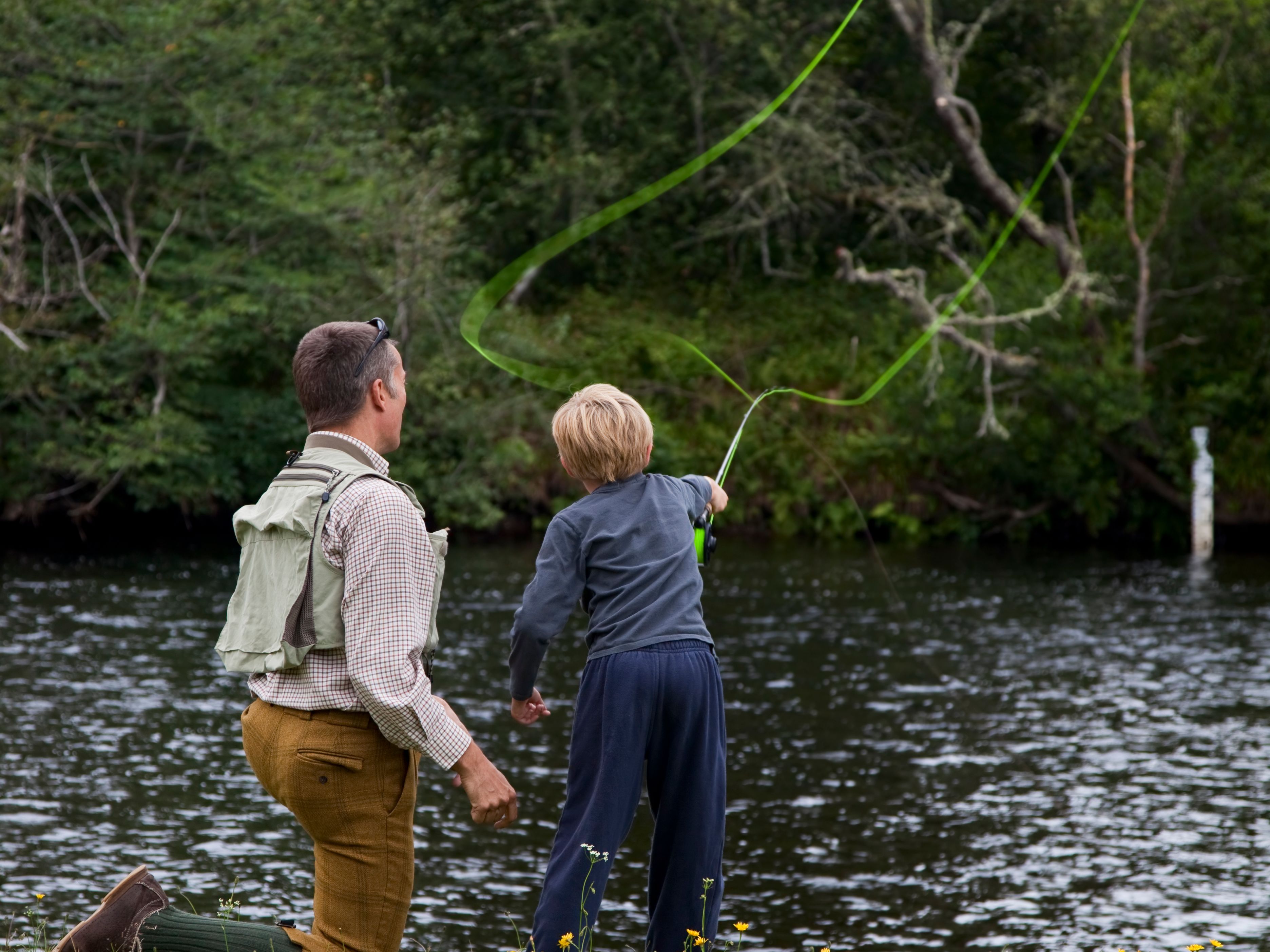 Adult and child fly fishing together by a riverbank with trees in the background