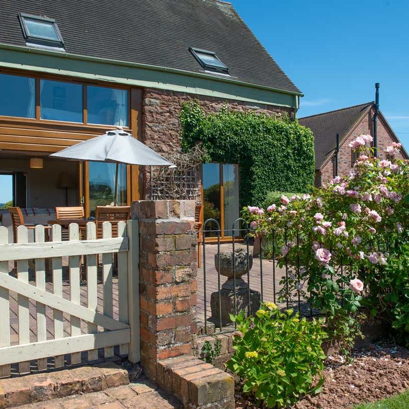 A charming cottage with a garden, white gate, patio furniture, and blooming pink flowers.