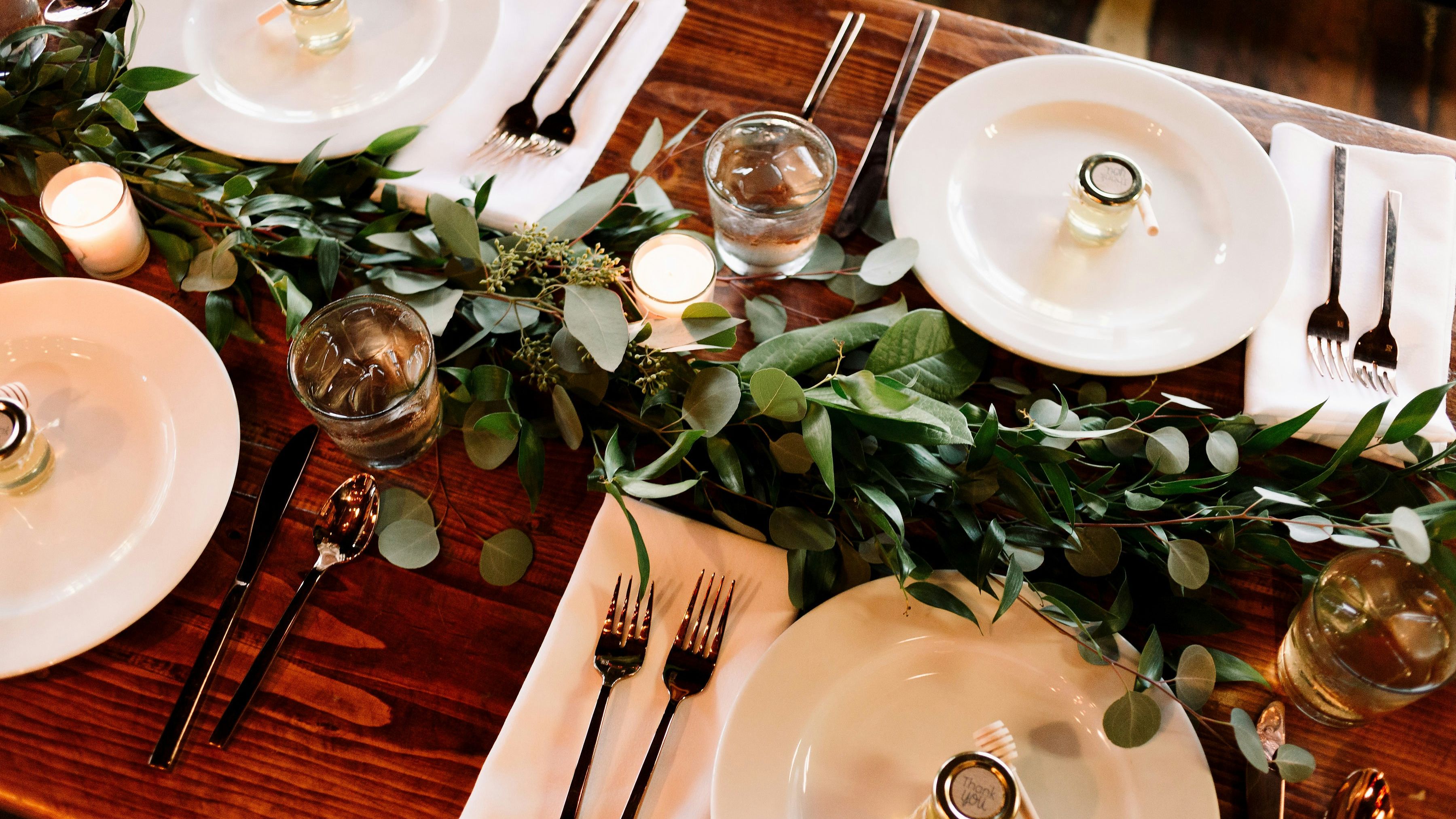 Rustic wooden dining table set with white plates, cutlery, water glasses, candles and a leafy green garland