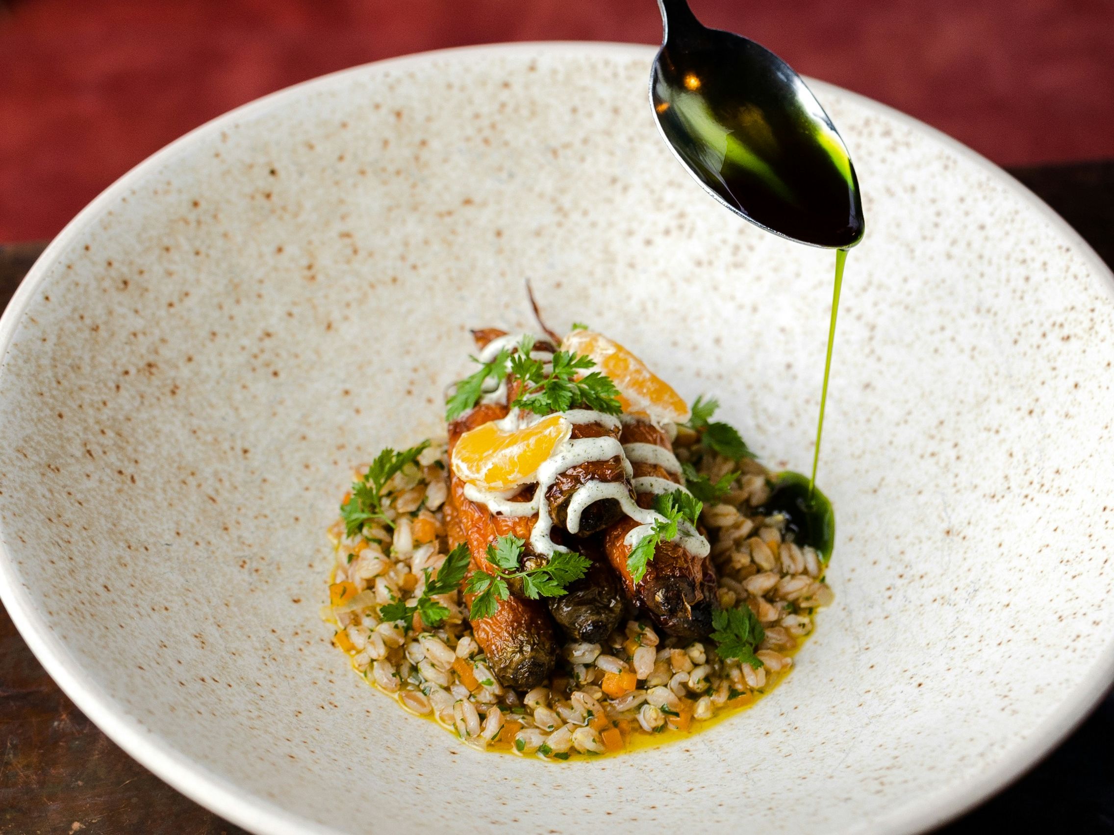 Hand drizzling green herb oil from a spoon over a plated dish in a speckled bowl