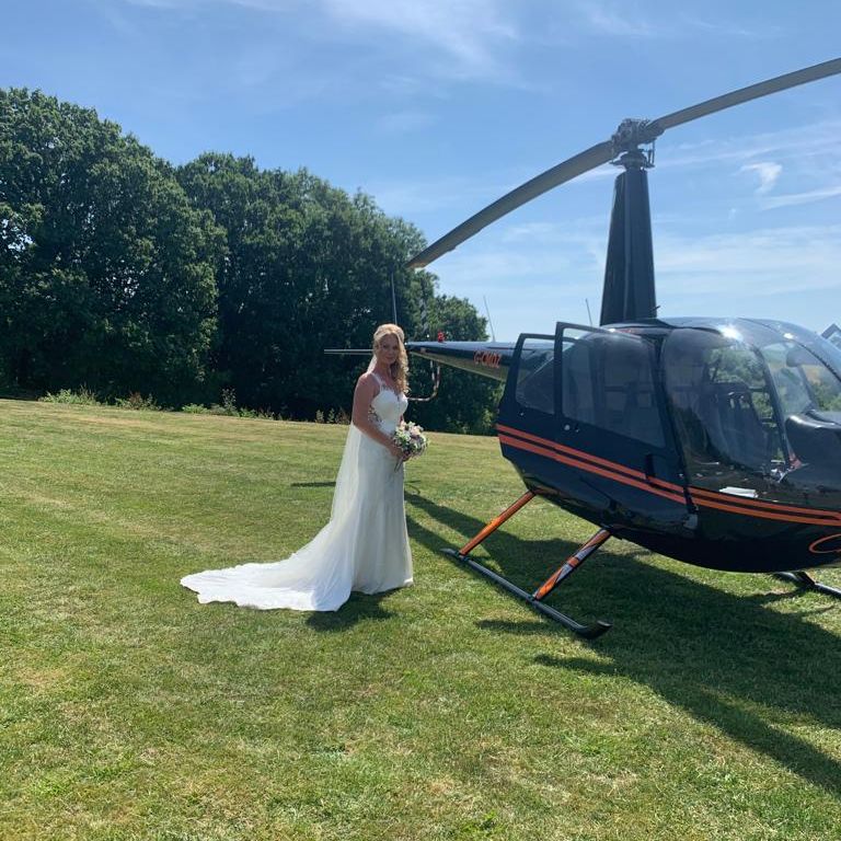 Bride in a white wedding dress holding a bouquet beside a black helicopter on a grassy field