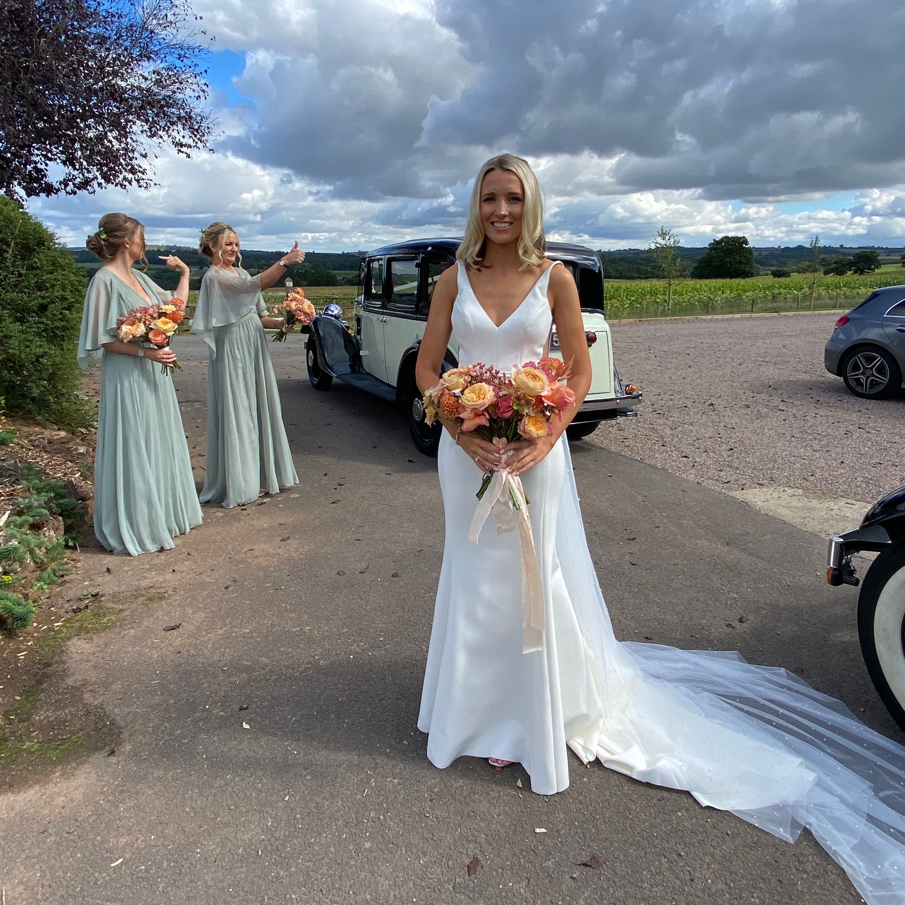 Bride holding a bouquet outside beside a vintage car, with two bridesmaids in pale green dresses in the background under a dramatic cloudy sky