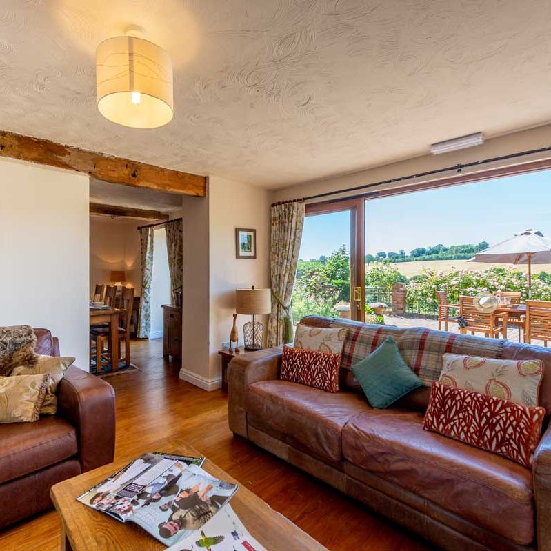 Cozy living room with brown leather sofas and large glass doors leading to a patio with outdoor seating and countryside view.