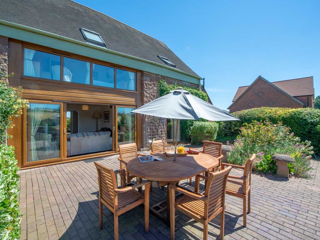 Outdoor patio with wooden table and chairs set under a large umbrella, next to a house with large glass doors and greenery.