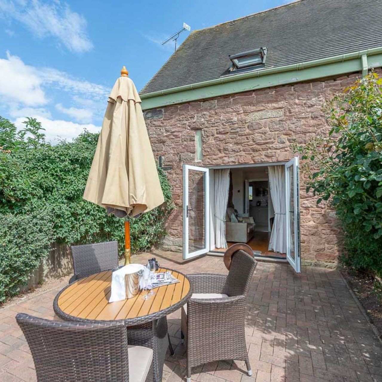 Outdoor patio area with round table, four chairs, parasol, and open doors leading into a stone house
