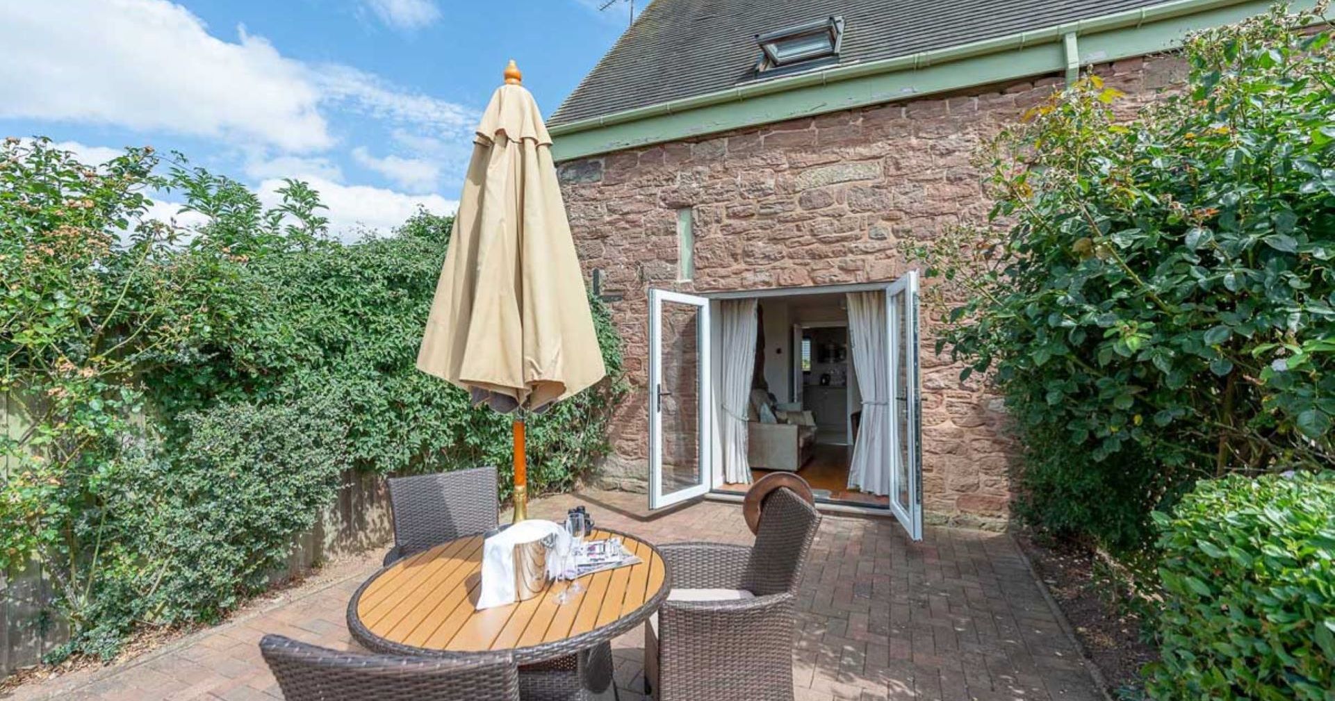Outdoor patio area with round table, four chairs, parasol, and open doors leading into a stone house