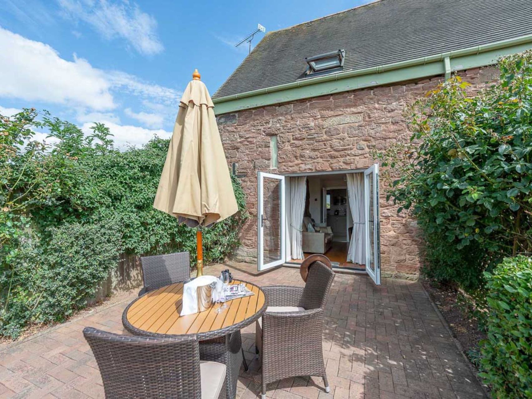 Outdoor patio area with round table, four chairs, parasol, and open doors leading into a stone house