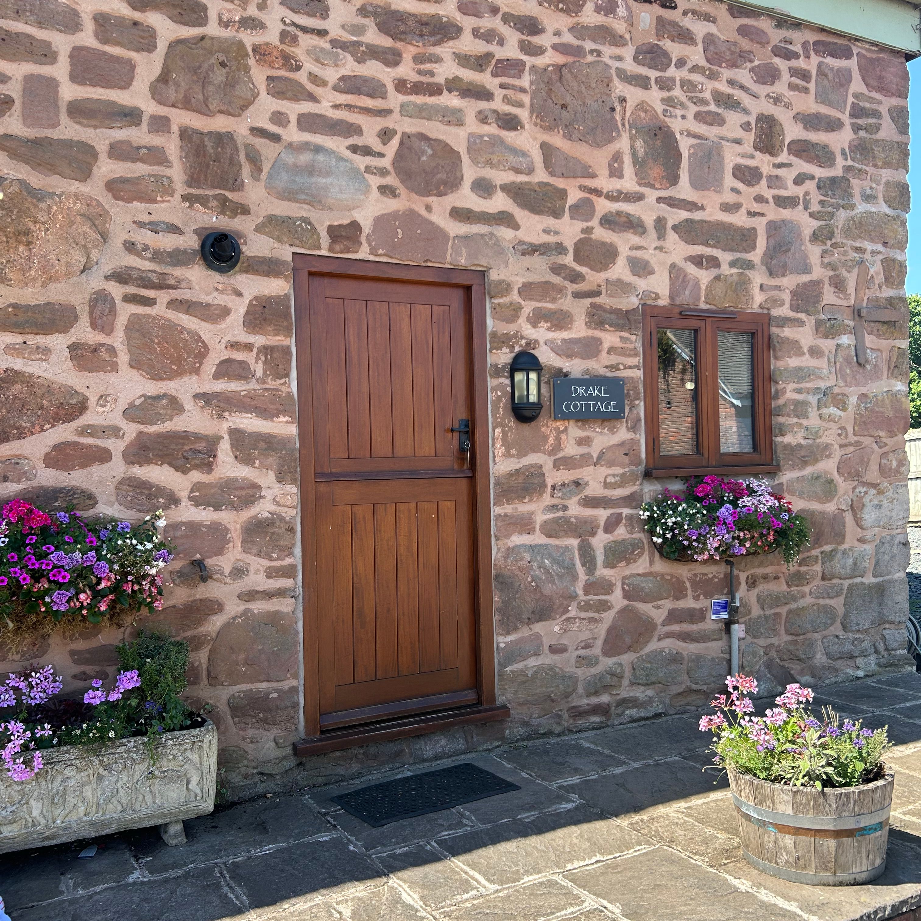 Entrance to a stone cottage with a wooden door, flower boxes, and a sign reading 'Drake Cottage.'
