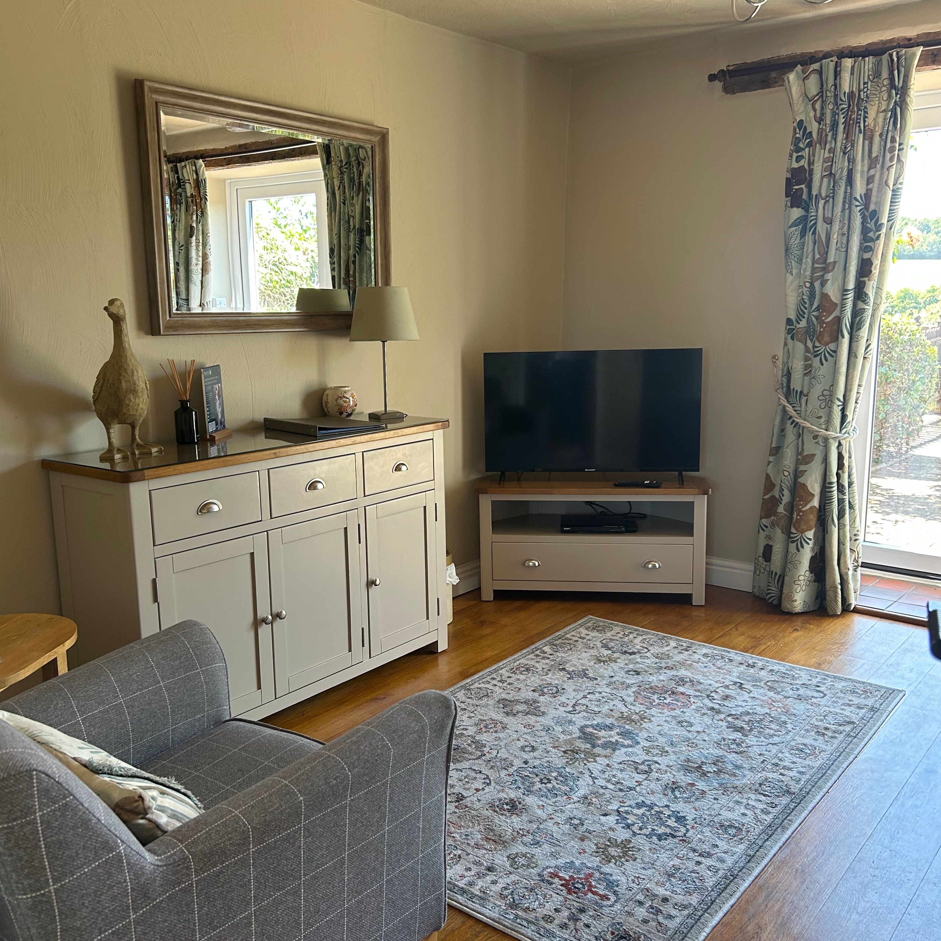 A cozy living room with a grey armchair, patterned rug, TV, sideboard, large wall mirror, and floral curtains.