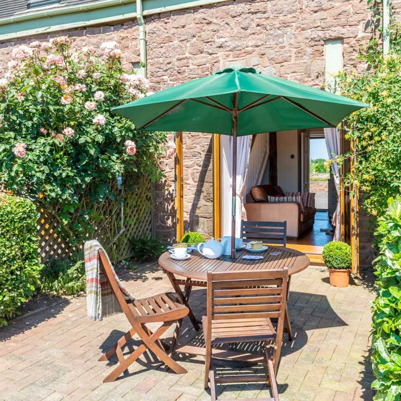 Outdoor patio with a round wooden table, chairs, and green umbrella beside a stone house
