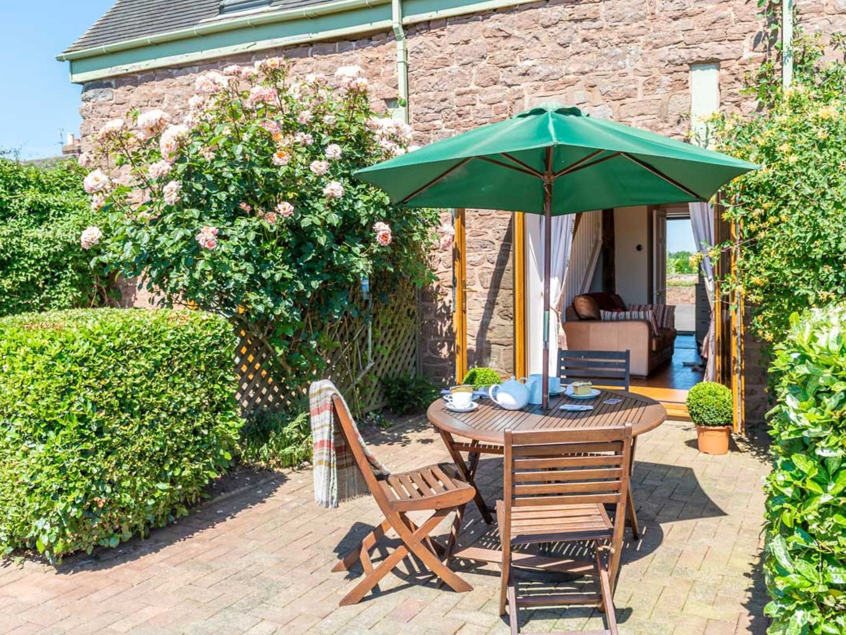 Outdoor patio with a round wooden table, chairs, and green umbrella beside a stone house