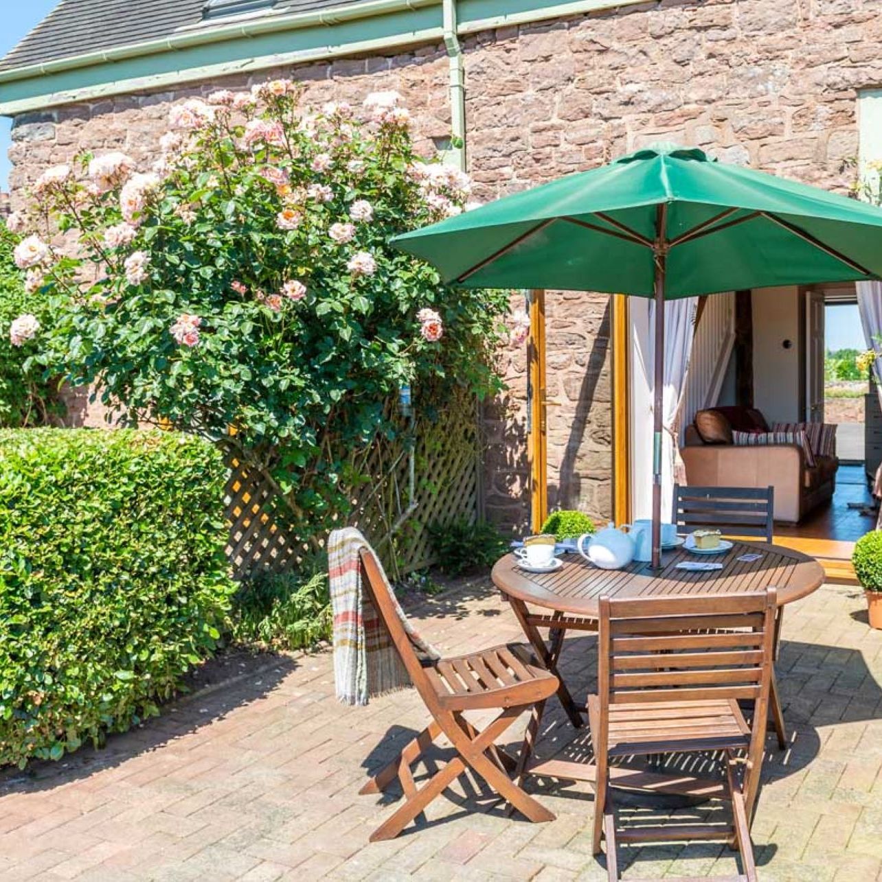 Outdoor patio with a round wooden table, chairs, and green umbrella beside a stone house