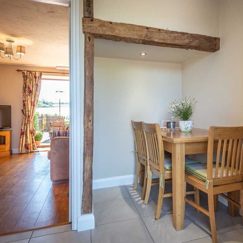 Cozy dining nook with wooden table and chairs next to a rustic beam, adjacent to a living area with wooden flooring and a TV.